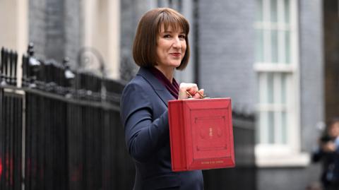 Chancellor of the Exchequer Rachel Reeves, who has short brown hair, wearing a navy suit with a burgundy top standing outside 11 Downing Street on Budget Day holding a red ministerial budget box/briefcase with golden calligraphy on the box and a gold handle.