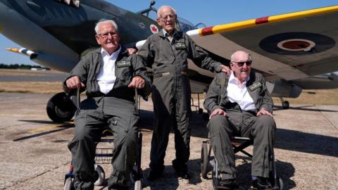 From left to right, Ivan Staines, 99, Paul Terry, 101, and Alfred Cooley, 100, pose in flying suits in front of a Spitfire at Biggin Hill Airport in Kent. Staines and Cooley are seated in wheelchairs while Terry stands between them with his hands resting on their shoulders. The aircraft is parked on the airfield behind them under a clear blue sky.