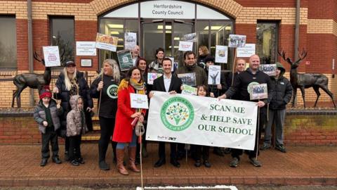 A group of adults and children posing outside the brick-built offices of Hertfordshire County Council in Stevenage. They are holding signs and a large white banner.