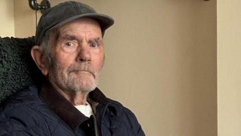 An elderly man wearing a dark grey cap looks towards the camera with a blank expression. He has grey stubble and is wearing a navy blue coat and white t-shirt.
