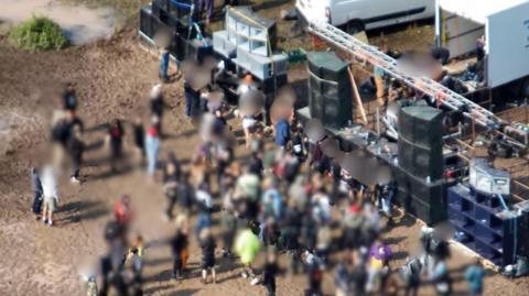 A crowd of about 60-70 people gathered around a makeshift stage made of stacked speakers. The ground is dusty and dirty, and everyone has their faces blurred out. There are large white vans parked in the background.