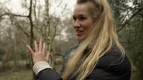A woman with blonde hair in a dark jacket gestures in the forest