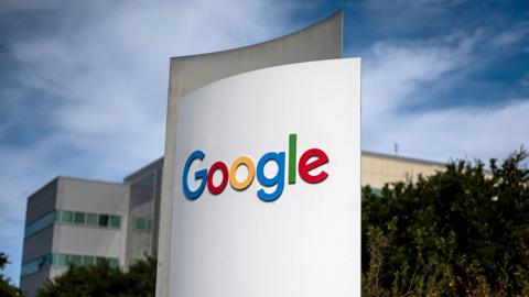 Google's multi-coloured logo is seen on a white sign in outside their headquarters, a grey and beige building with trees in the foreground and wispy clouds against a blue sky, in Mountain View, California in 2024