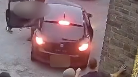 A black with bright red reverse lights on. The passenger-side door is being thrown open. A group of people watch from behind
