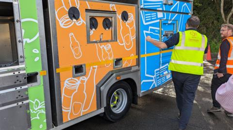 Two men wearing hi-vis waistcoats next to a recycling truck. One is holding a bag, while the other is putting something in the van.