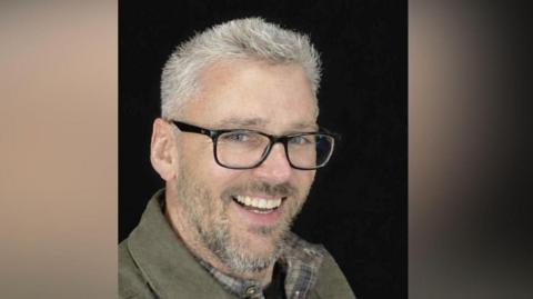Craig Hurcombe smiles at the camera. He has short grey hair and is wearing black, thick-rimmed glasses and a brown shirt. He is pictured against a black background.