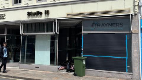 The front of a dry cleaners which has been boarded up after a fire. The dry cleaners is called Rayners and is next to a hair salon. A green wheelie bin is outside the shop. A boy wearing a school uniform is walking along the street.