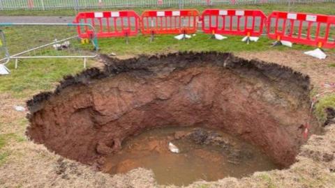 The large, round sinkhole is full of mud and water. There is a line of low plastic red fencing behind it with taller metal fences to the side.