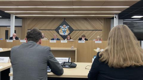 Councillors at the cabinet meeting of Stockton Council. The image is taken from the public gallery, with the back of two council officers - a man and a woman - in the foreground. The cabinet is sat opposite them at a long raised table, with their names displayed on a sing in front of each. Behind them is the emblem of the council, displayed on a herringbone patterned wooden panel. 