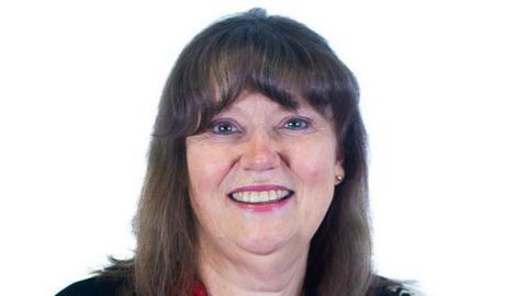 Cate Caruth is smiling at the camera in front of a white background. She has shoulder length brown hair with a fringe. She is wearing a dark coloured top.