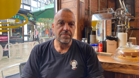 Pete Masters, wearing a blue tshirt, sat in front of the wooden counter of his cafe in Hereford, with a window showing the street behind him