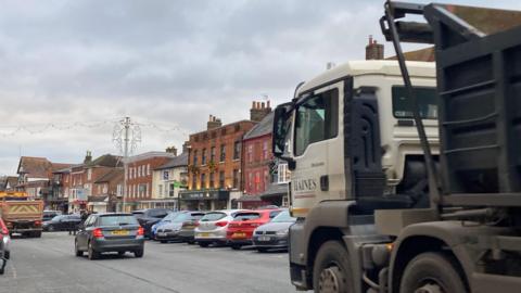 Marlborough High Street with the parking in it full and a truck driving through behind a car and another heavy vehicle.