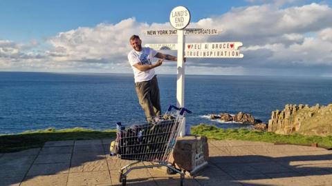 Perry Pel Scopes stands on Bruce the shopping trolley in front of the Land's End sign. The sea is in the background and the sky is blue. He is wearing a white tshirt and brown trousers. The sign says 'Pel & Bruce a true inspiration'