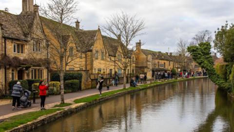 Images of tourists stood near water talking pictures in Bourton-on-the-Water. There are yellow-stone houses in the background near trees.