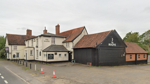 The Magpie Inn is seen in the picture. The building spans across the whole picture, with the main part of the pub situated on the left, which is a white building with signs hanging up. 