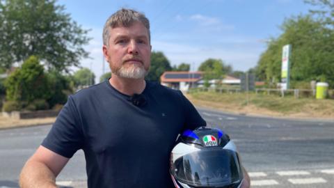 Lee is standing with a a navy t-shirt holding his motorcycle helmet on his right hand side. He has short grey hair and trimmed grey beard
