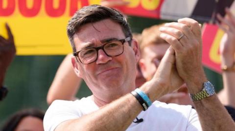 Andy Burnham is pictured wearing a white T-shirt and clapping his hands, with Labour supporters in the background.
