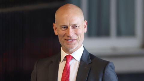 A bald man in a dark suit, white shirt and red tie.