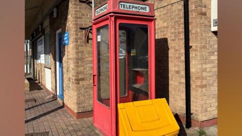 A traditional red telephone box positioned on a brick-paved walkway beside a building made of tan-colored bricks. The telephone box has glass panels on all sides, and the word “TELEPHONE” appears in white lettering on a sign at the top of the box on at least two visible sides. The paint on the box appears worn in some areas.