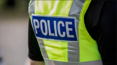 A general image of the back of an unidentified police officer. They are wearing a black shirt. On top they are wearing a high-vis green vest with a blue badge in the centre which reads 'police' in white writing.