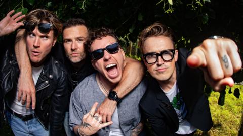 Four members of McFly are posing. Three of them are wearing glasses, one member on the right is pointing at directly the camera.