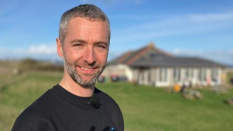 A man stands in front of a cafe building on the coast in Devon. It is sunny, with people sitting at tables. The man wears a black top and is smiling.  