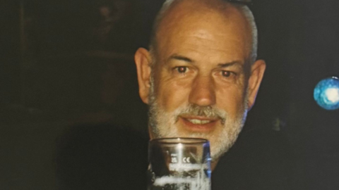 A man with a pint glass in front of him. He has short grey hair and a grey beard.
