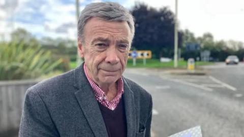 A man with grey hair and a blue jacket over a blue pullover and a red checked shirt, standing beside a road