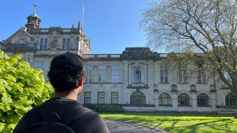 Jordan looks towards the main Cardiff University building, there are green bushes on his left and grass and a tree on his right. The building is ornate and have lots of arches and intricate details. Jordan has thick curly hair and is wearing a black cap, jumper and rucksack. 