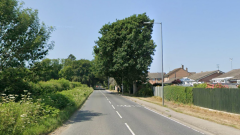 A Google Maps picture of Warmwell Road, a road with houses on the right hand side and a grassy area on the left.