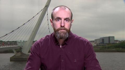 Mark Durkan - a man with receding hairline and a brown beard streaked with grey. He is wearing a plum coloured shirt and is sitting against a background of the peace bridge in Londonderry.