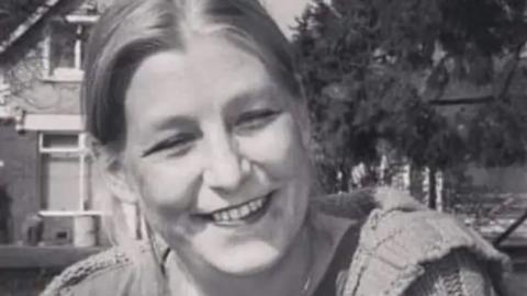 A black and white picture of Dawn Sturgess smiling at the camera, wearing a t-shirt, cardigan and a necklace. She is sitting on the grass in front of a house and a tree.