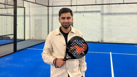 A man in a cream overshirt holding a black padel racket is smiling on an indoor padel court