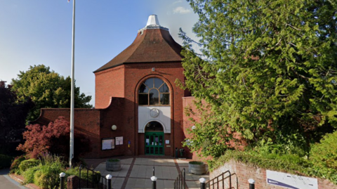 A Google Street image of red-bricked building which you enter through a main atrium. It is down a set of stairs and there is a flag pole on the left.