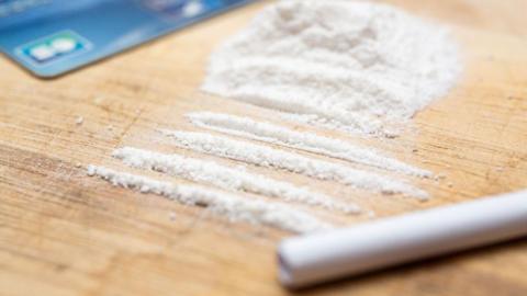Illustration photograph of drugs imitating cocaine on a board in lines or traces with a straw in the foreground and a credit card in the background.