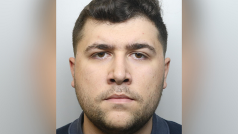 A close-up, front-facing custody photo of a man with short dark hair and facial hair, wearing a dark shirt. He has a neutral expression and is positioned against a plain, light-coloured background.