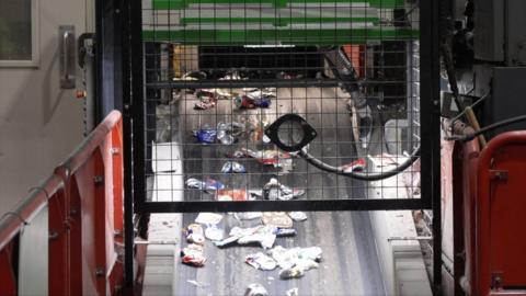 Rubbish such as cans on a grey conveyor belt. Above there is a black fence