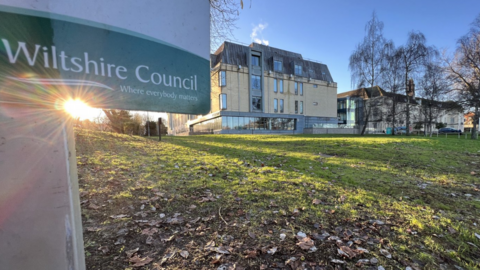 A picture of Wiltshire Council's headquarters in Swindon, as taken from afar. The building is standing in the middle of a large grassy area, and is a sand coloured building with lots of windows. In the foreground there is a Wiltshire Council sign. 