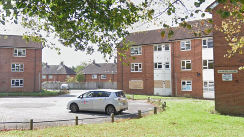 Several windows are visible in a three-storey building on the right and three sets of windows can be seen in a three-storey building on the far left. A car is parked in the foreground.