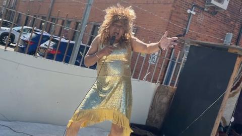Man in a gold glittery dress with a mircophone singing on a stage outside his shop. 