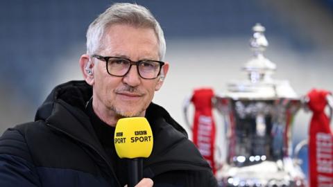 Gary Lineker in a thick coat holding up a yellow BBC Sport microphone, with the FA Cup next to him in 2021