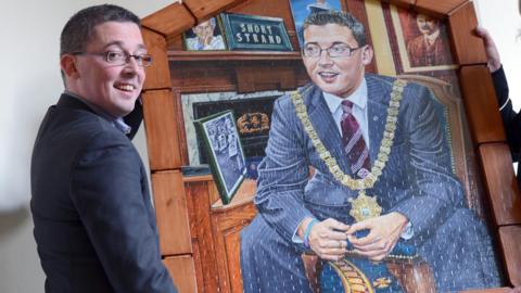 Niall Ó Donnghaile carrying his portrait. He is wearing a navy suit, blue shirt and red jumper. 