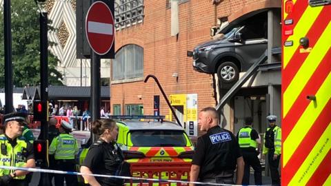 A grey car pokes out from a first storey window with its wheels hanging over the street, as emergency services look on.