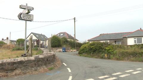 A wide image of the junction into Llangwnnadl village. On the left there are road signs attached to a pole. On the right of the image are houses. 
