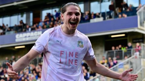 Lorent Tolaj, in action for Plymouth Argyle, with his arms out wide and smiling celebrating scoring a goal against AFC Wimbledon.