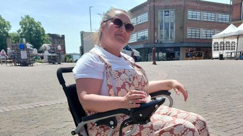 A brown haired woman wearing sunglasses and a white t-shirt with pink dungarees in a black motorised wheelchair.