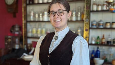 A woman wearing glasses dressed in Victorian period costume stands in front of rows of bottles.