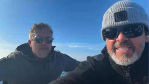 Two men sat next to each other on a boat. Matthew Parker (right) is wearing a grey beanie hat, black coat and black sunglasses. He is sat next to Mike Bates who is wearing a black coat and sunglasses. 