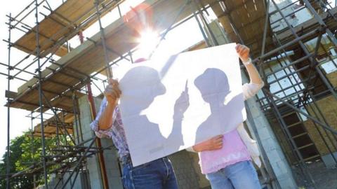 A couple stand with their faces obscured but silhouetted by a large piece of white paper, as they stand on a building site