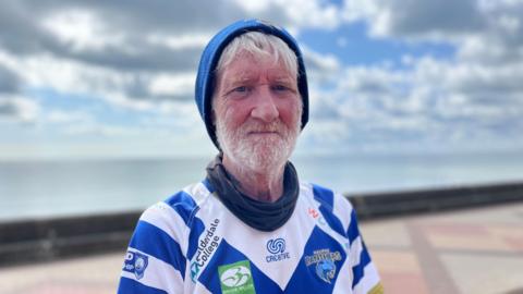 An older white man looks directly into the camera. He has a serious expression. He has white hair, a white beard, and blue eyes. Two small tattoos on his neck can just be seen. He is wearing a blue woolly hat and a blue and white sports shirt with a 'Halifax Panthers' logo. He also has a black roll-neck. The background is blurred but still visible - the seafront can be seen. 